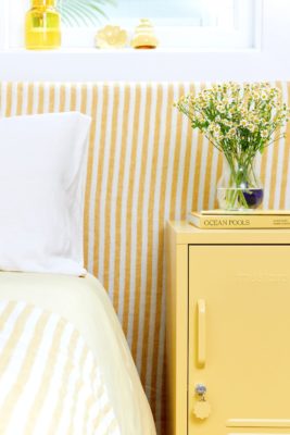 Bedroom with yellow and white striped headboard, matching bedsheets, and a butter yellow metal locker acting as a bedside table.