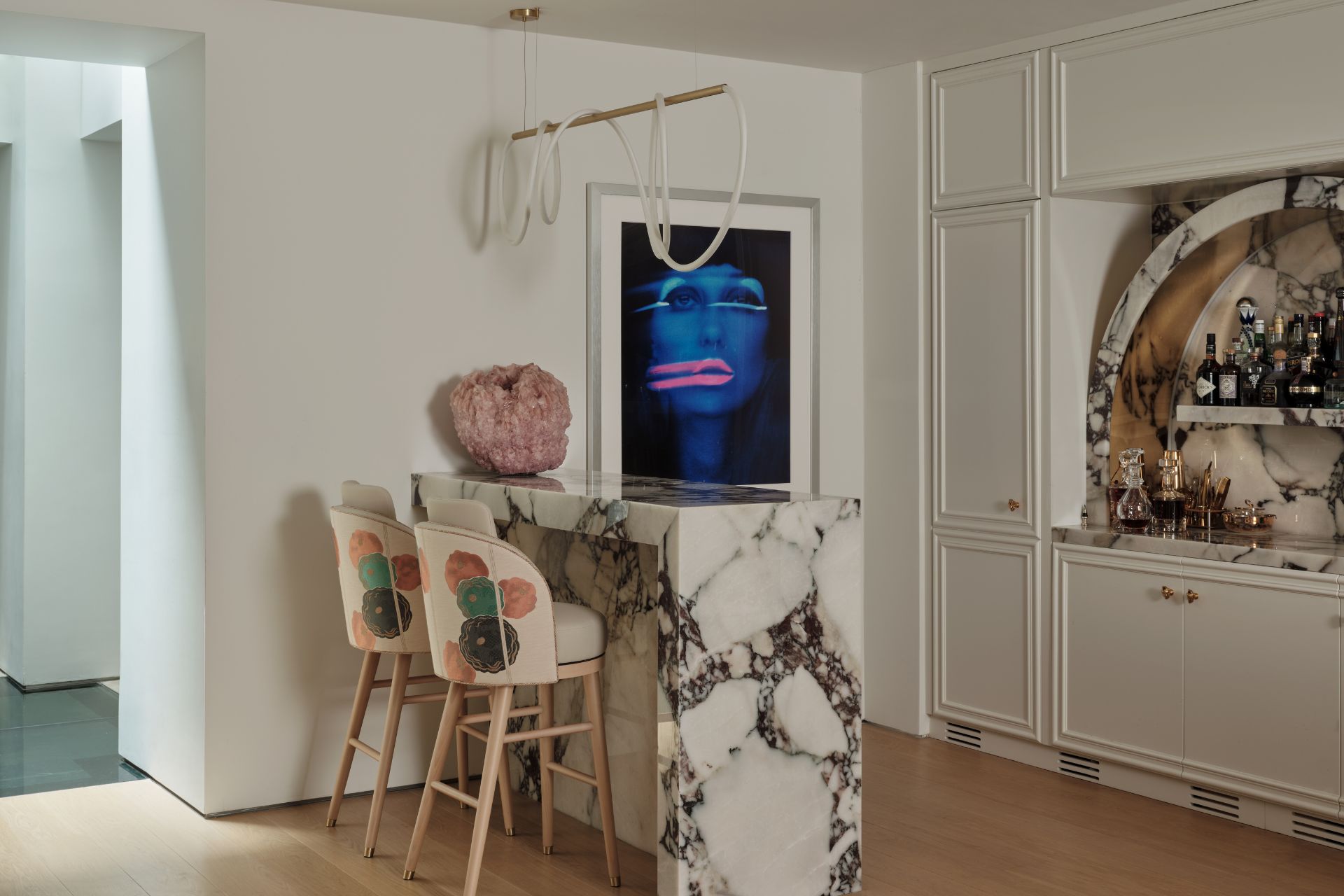 Kitchen with pale wooden floors, a white and grey marble breakfast bar, floral applique bar stools and a pink crystal vase.
