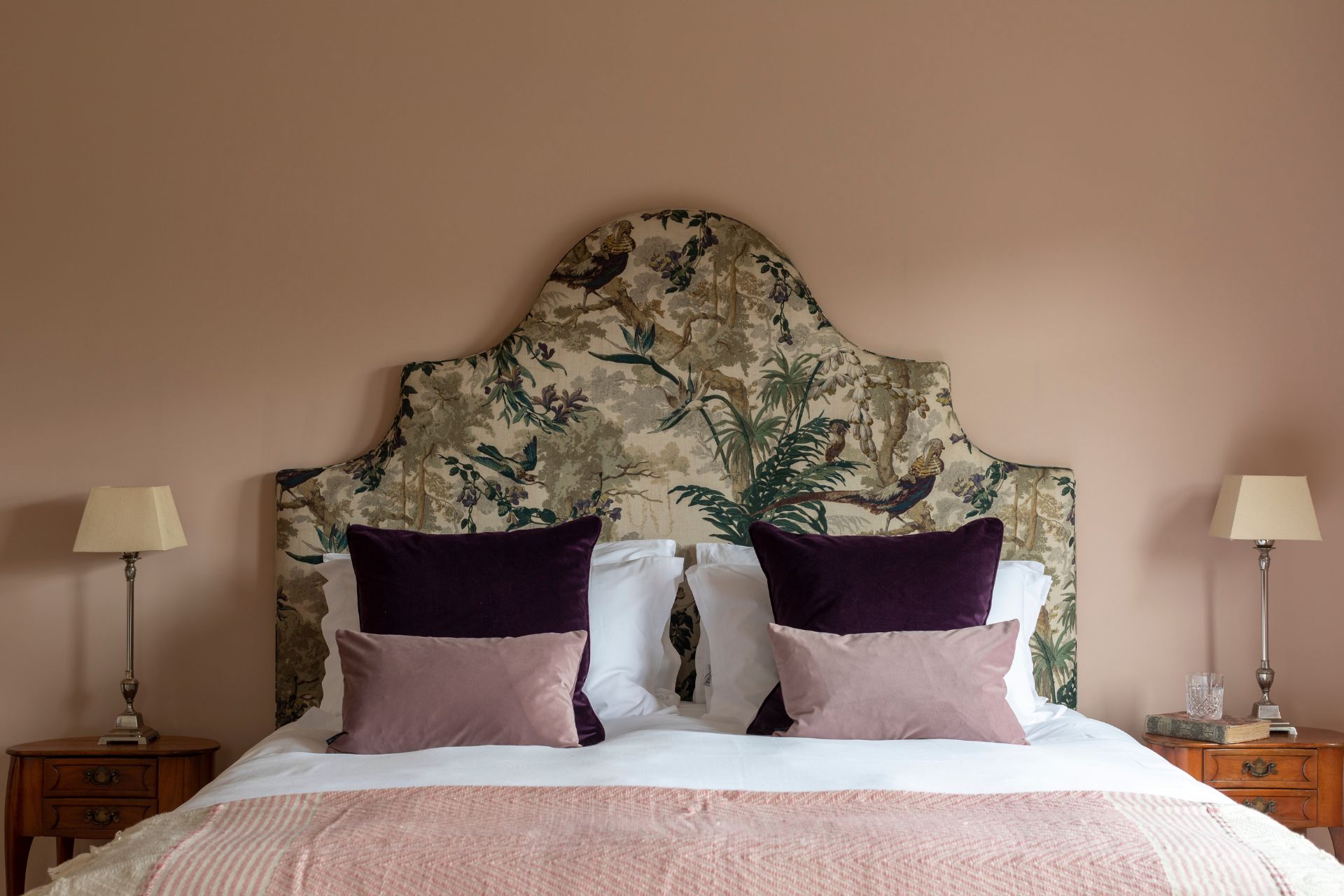 Bedroom with green botanical print headboard, a pale pink bedspread, and pale pink and black cushions.