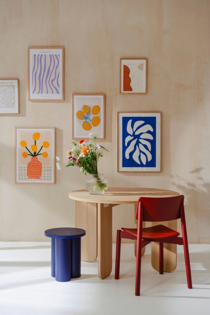 Dining area with whitewashed walls, colourful prints, a circular wooden table, a red chair and a blue stool.