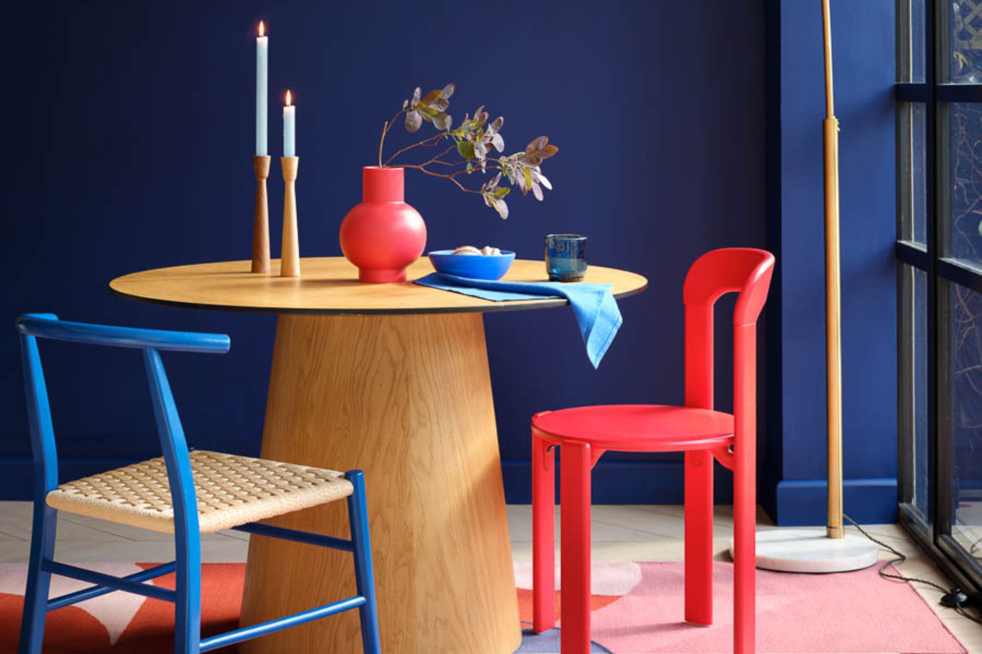 Navy dining room with pale wooden circular table, a red chair, and a blue chair with a rattan base.