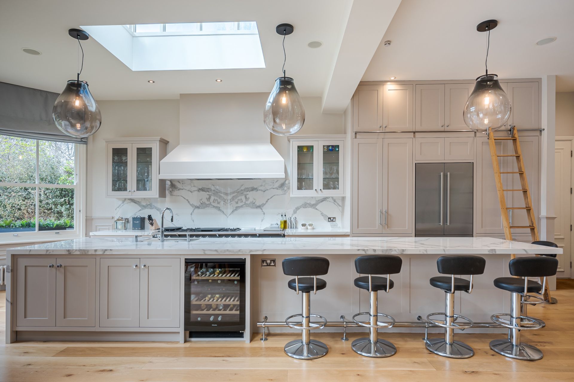 Modern kitchen with white cabinets, white marble worktops anda large island with black bar stools and a built-in wine fridge.