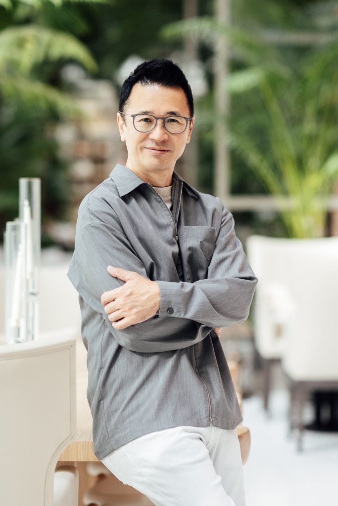 Clint Nagata standing in a garden earing a grey linen shirt and white trousers