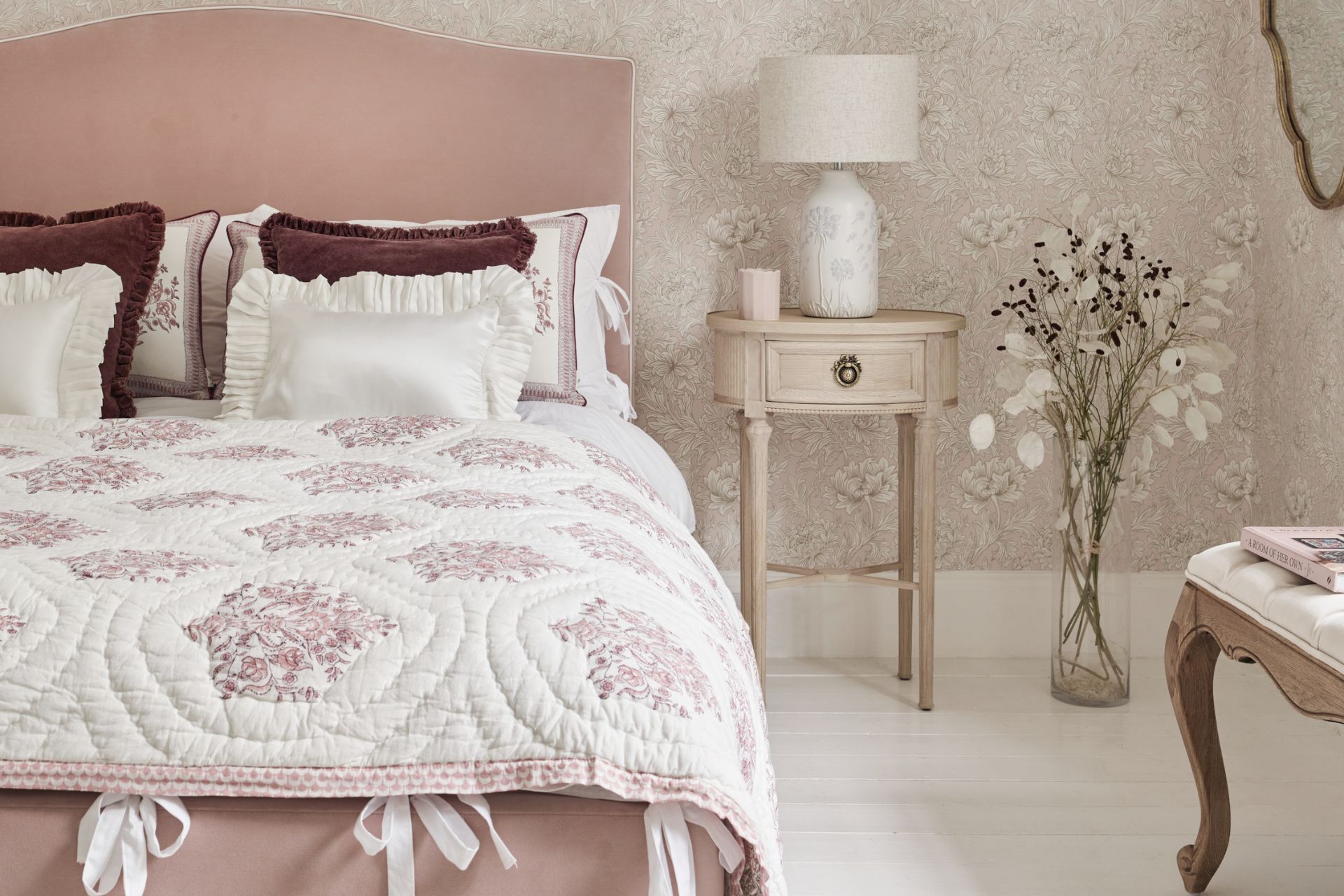 Bedroom with pale pink velvet headboard, pink floral bedding with white ribbons, and floral wallpapet