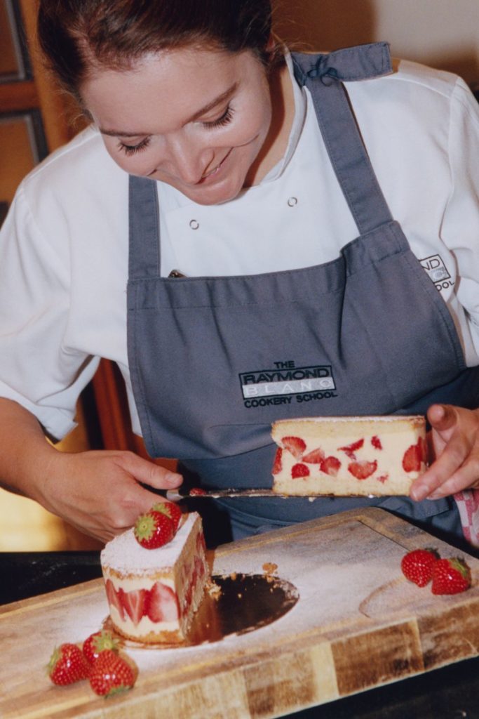 Chef wearing grey Raymond Blanc apron, serving slices of a strawberry cheesecake