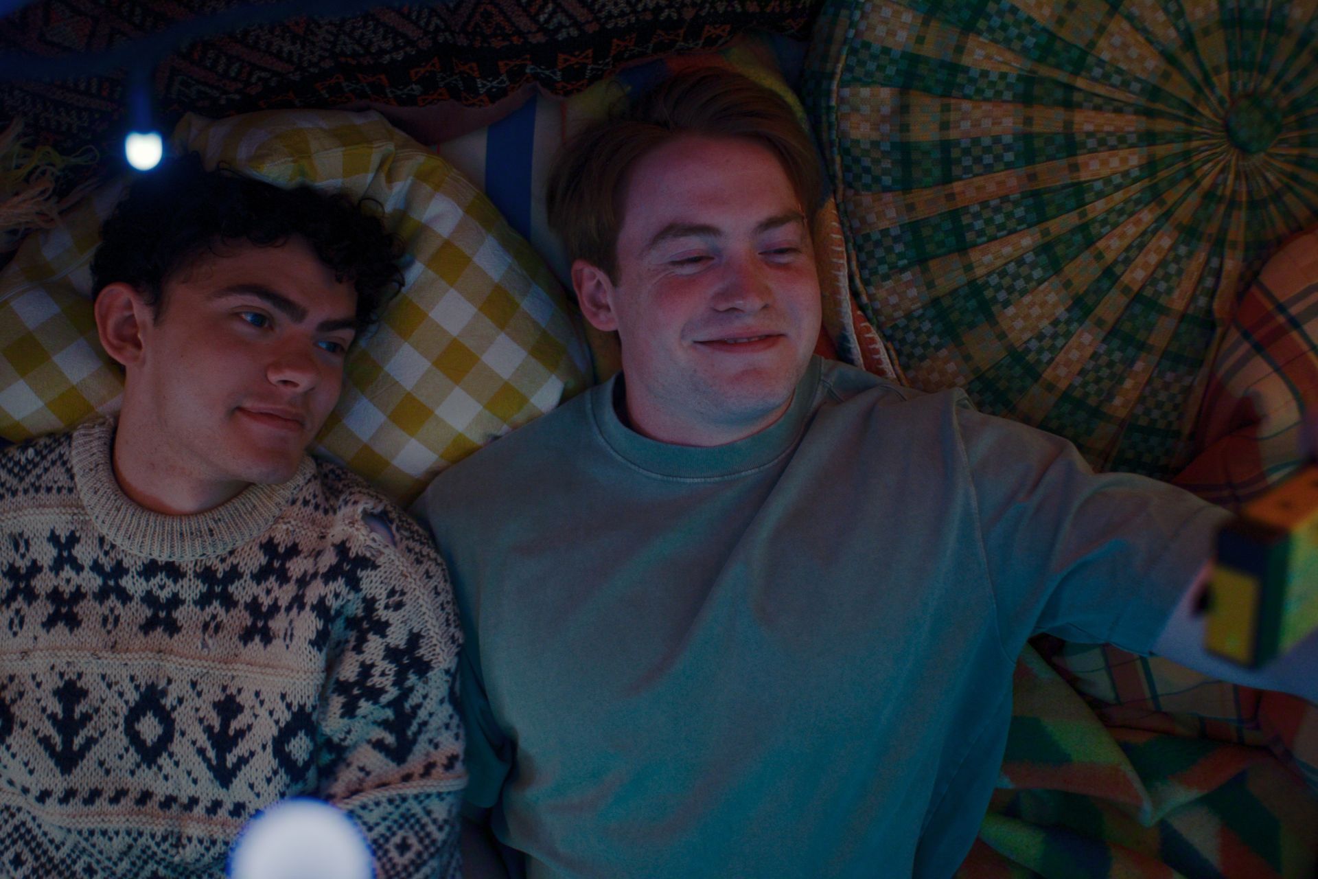 Two characters in Heartstopper lying in bed and smiling up at a disposable camera.