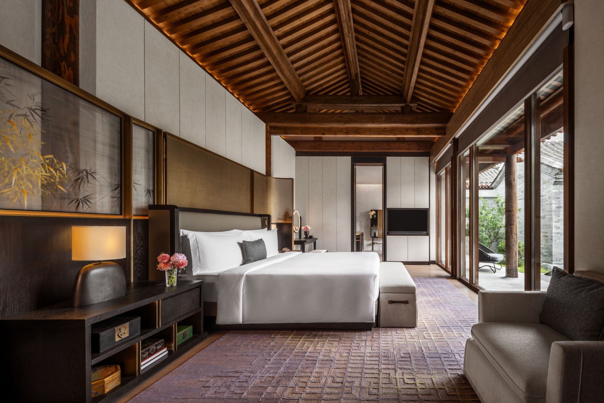 Bedroom with wooden ceiling beams, white bedding, a grey armchair, and floor to ceiling windows overlooking a courtyard