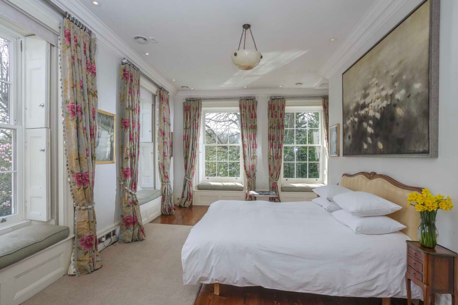 Bedroom with large French windows, floral curtains, white bedding and a painting above the bed