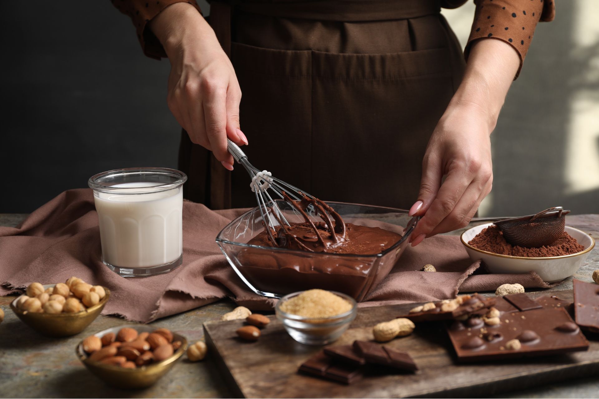 a person's hands mixing a bowl of melted milk chocolate. there are also bowls of nuts and a glass of milk on the table and a chocolate bar