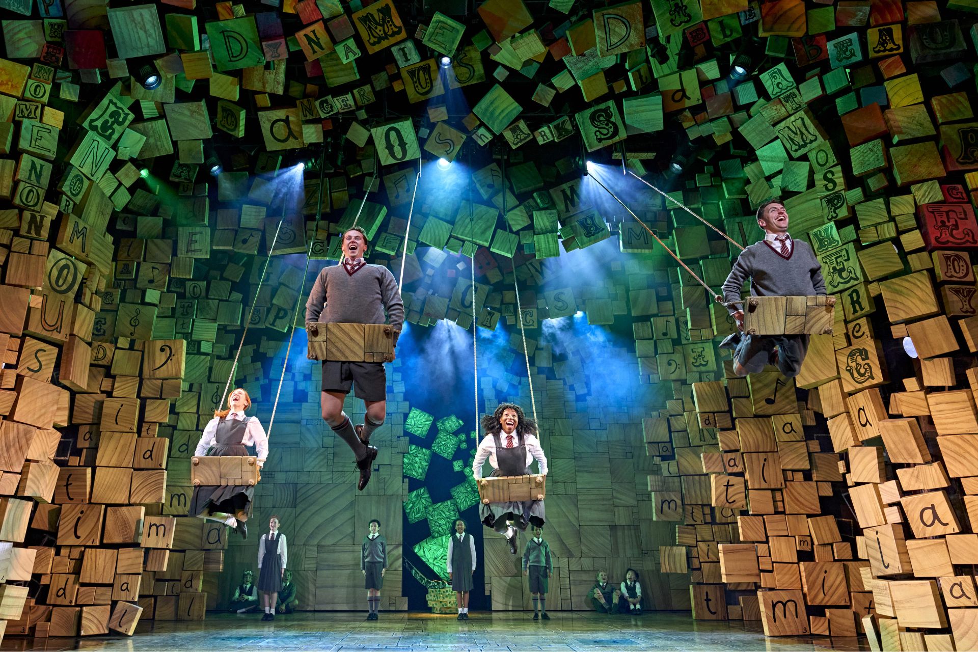 four actors dressed as school children lay stomach-down on giant swings in the air on a large stage