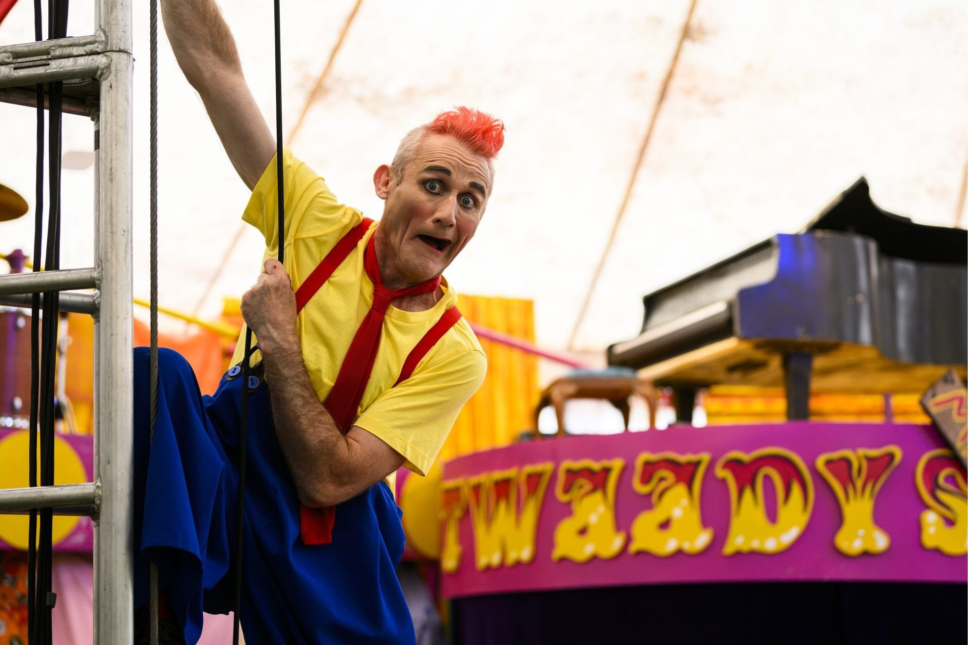 a clown with a red quiff is dressed in a yellow t shirt, red tie and blue trousers and hangs off a rope