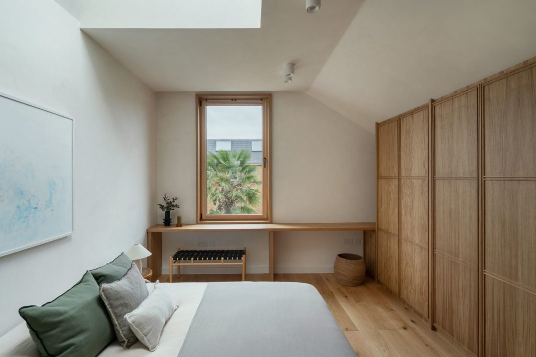 Japandi-style bedroom with oak fitted wardrobes, grey bedding, and a wood-framed sash window.