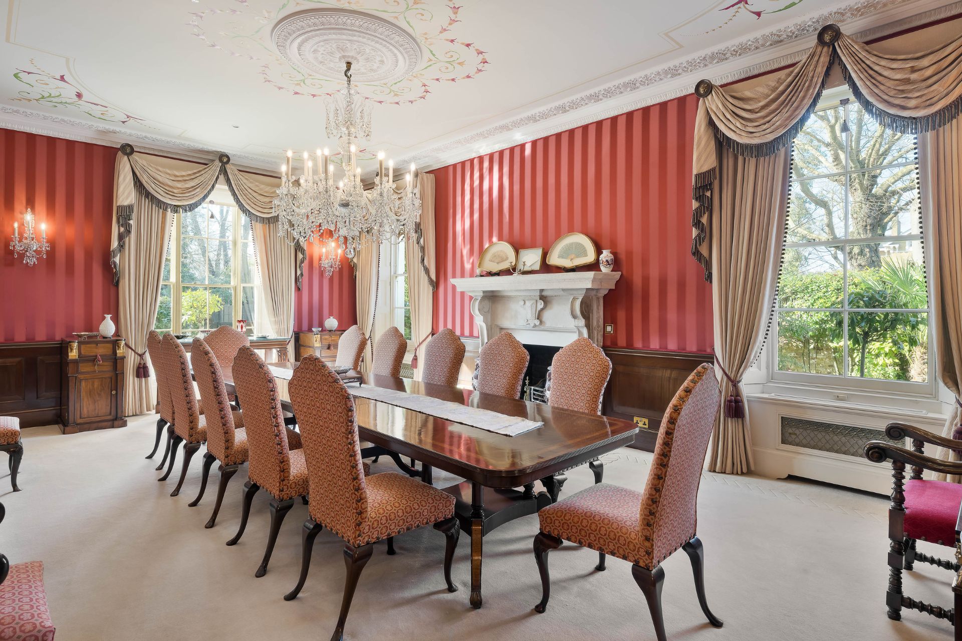 Dining room with wooden table, red velvet chairs, red striped wallpaper and a chandelier.