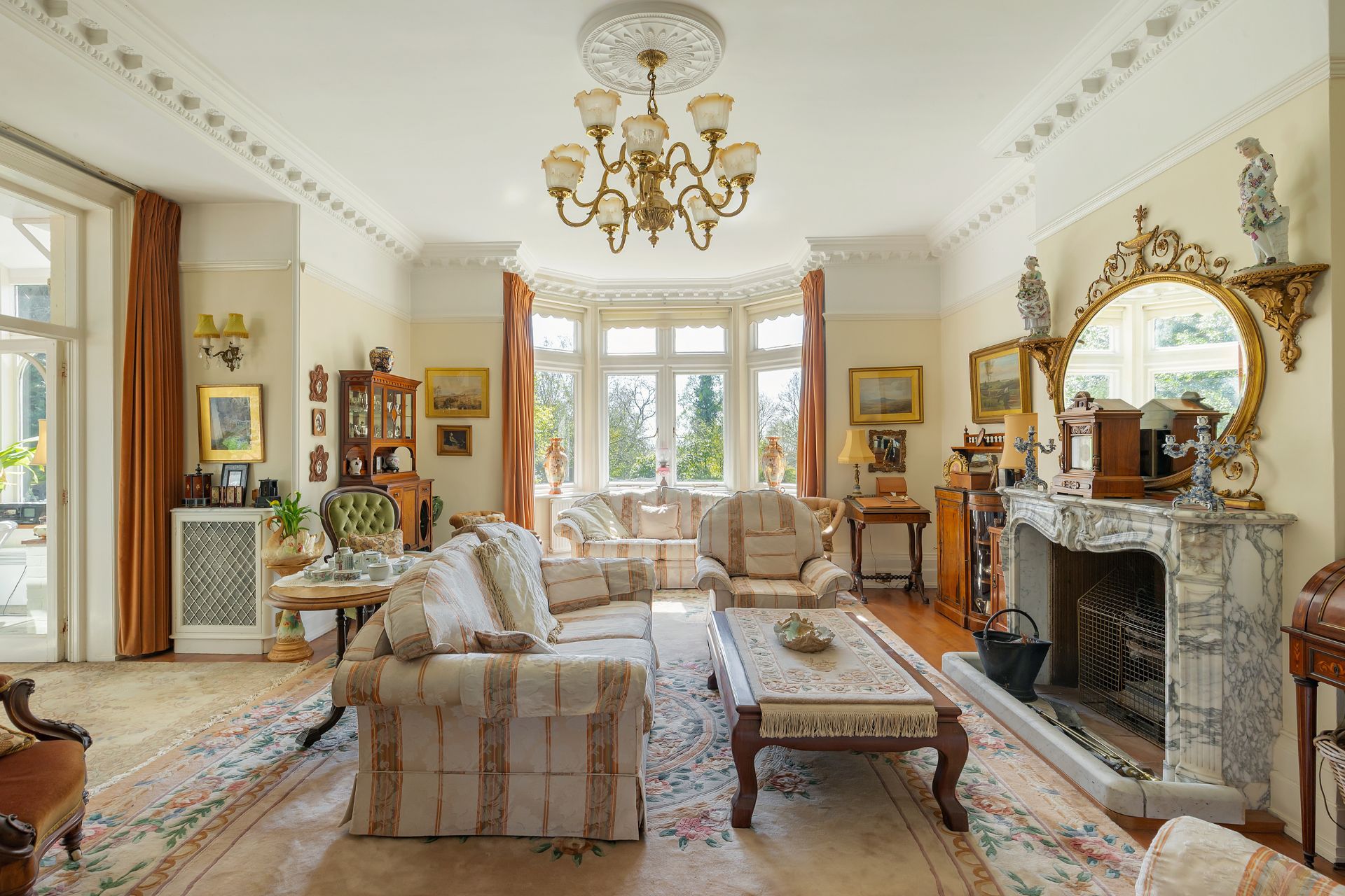 Living room with bay window, cream and golf sofas, a marble fireplace, and a chandelier.