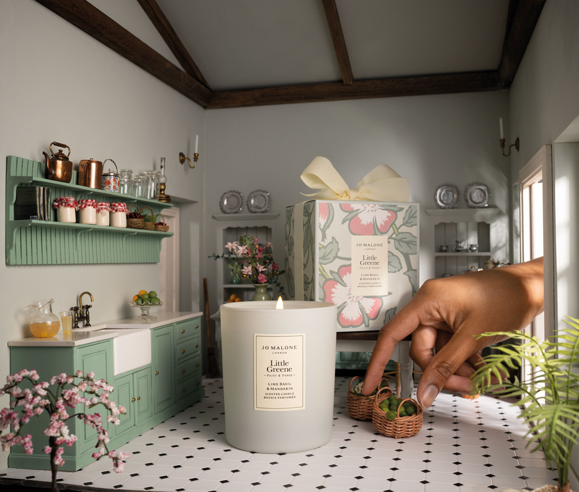 The Lime Basil and Mandarin candle in a doll house kitchen. A hand is coming through the window