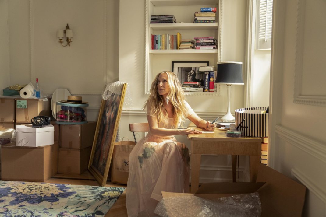 Sarah Jessica Parker as Carrie Bradshaw sitting in a new apartment with a bookshelves, rug and cardboard boxes