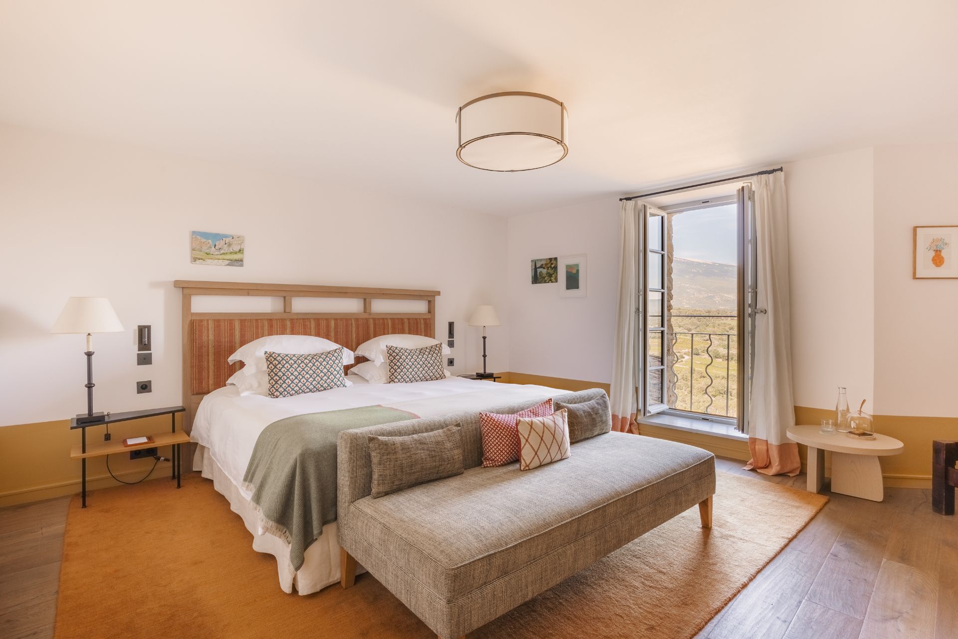 Large hotel bedroom with wooden bedrframe, white bedding, a sage green bedspread and a mustard yellow rug, with views of Provence