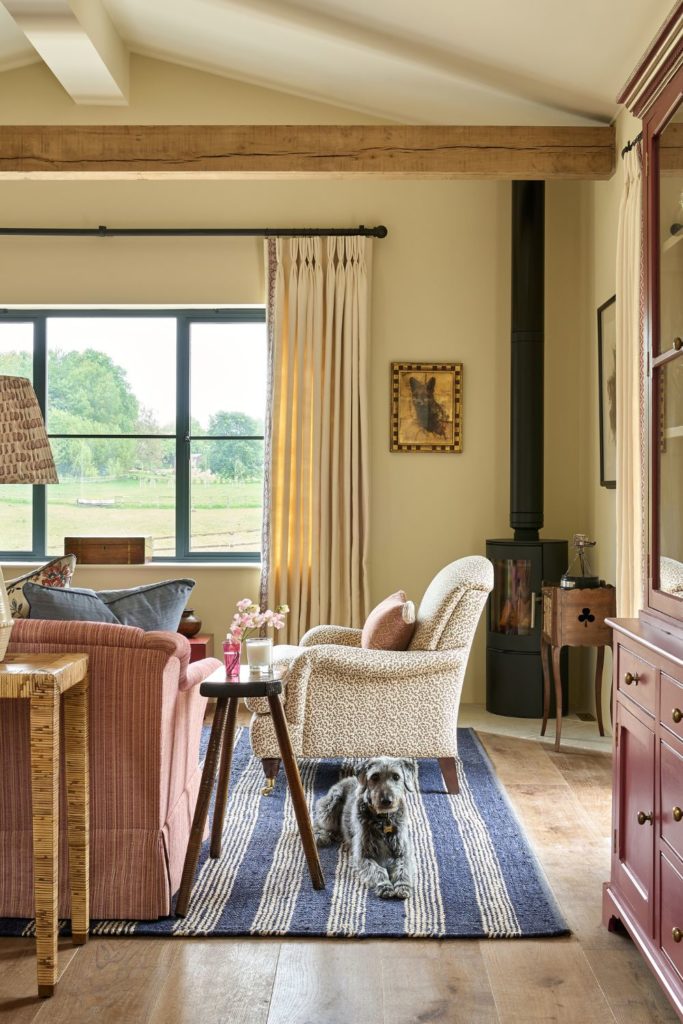 Barn conversion living area with a cream patterned armchair, blue and white striped rug, pale pink sofa and a woodburning stove