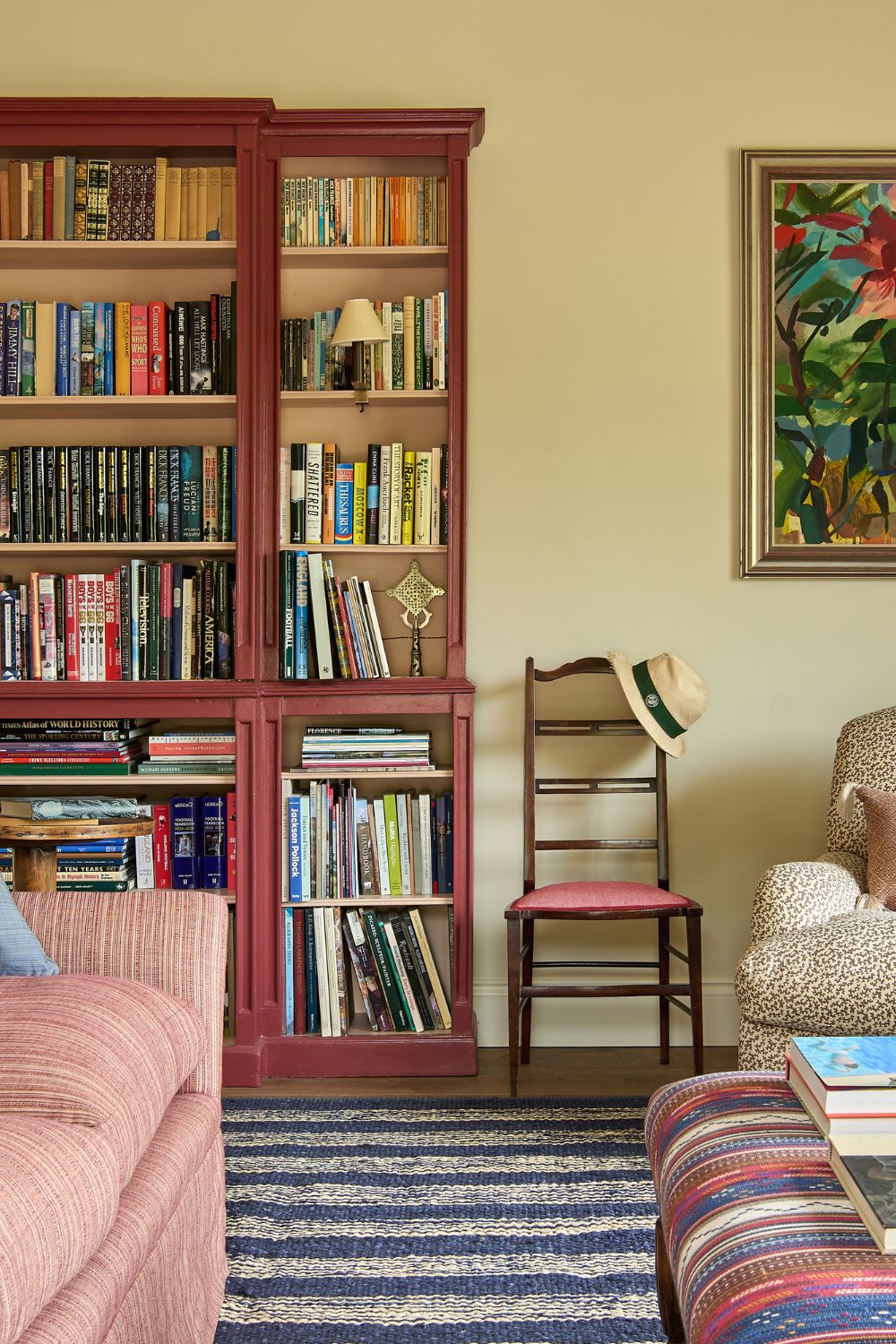 Living room with a blue and white striped rug, a pale pink sofa, a red and blue striped ottoman and a plum-coloured bookshelf