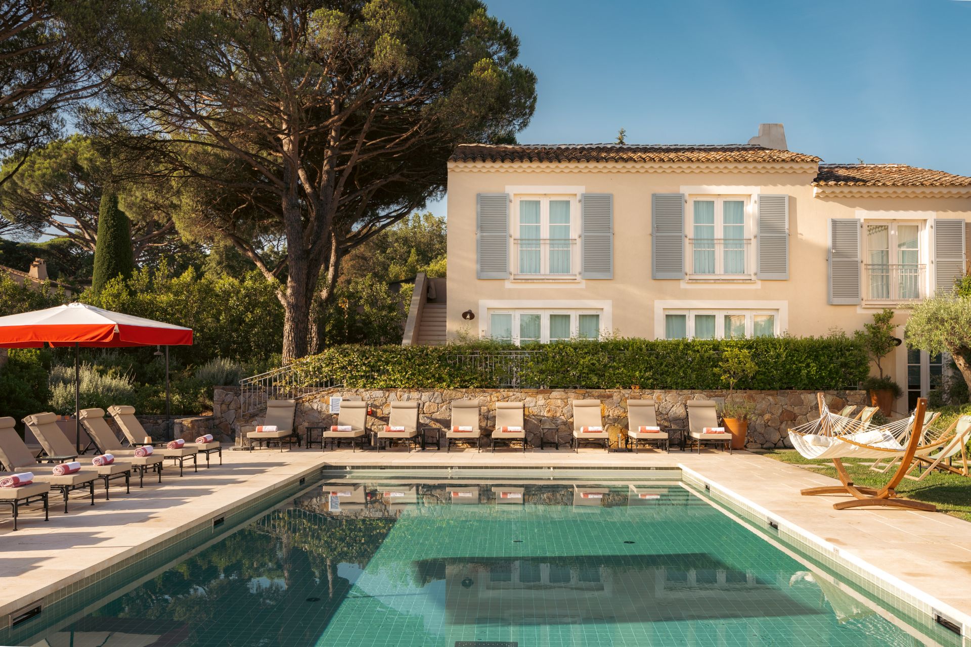 Hotel pool bordered by hammocks and red and white parasols, with a stone building behind