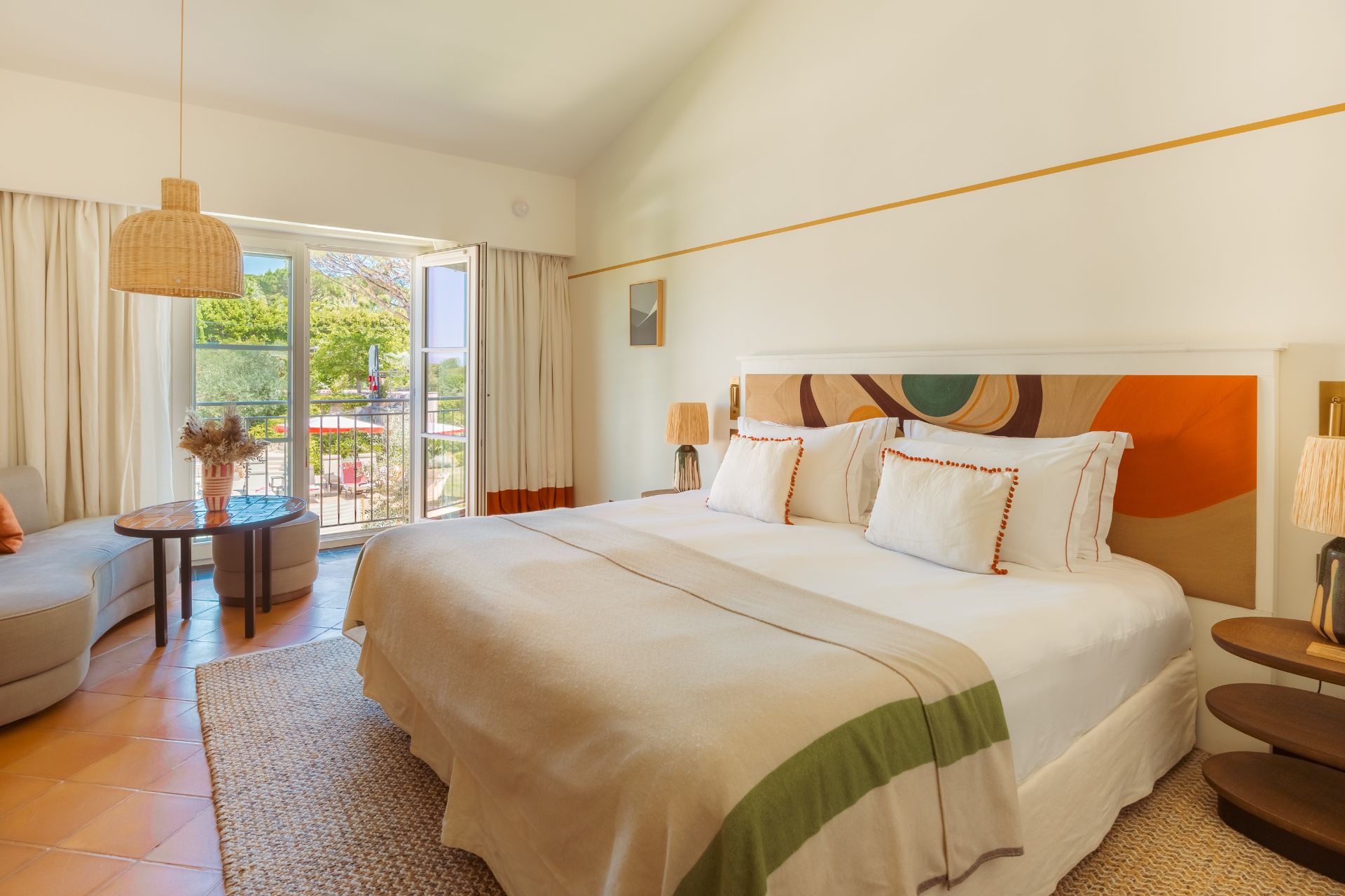 Bedroom with abstract tapestry headboard, wicker lampshade, red-trim cushions and a Juliet balcony overlooking a swimming pool