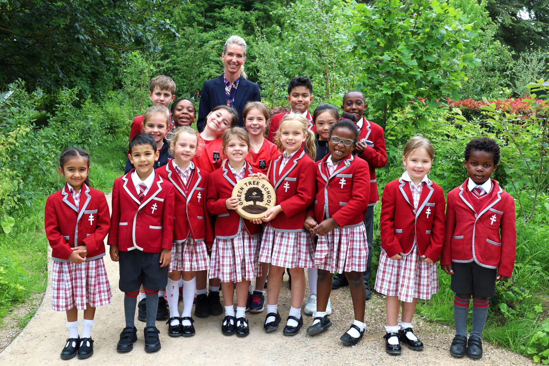 Young pupils in red uniforms stand outside with their teacher holding a plaque