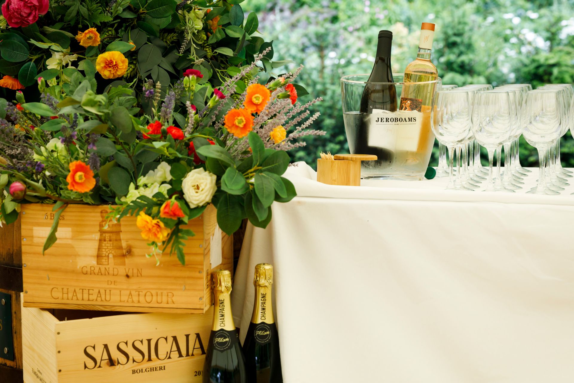 White table with champagne bottle and glasses next to a flower display and wooden crates of wine