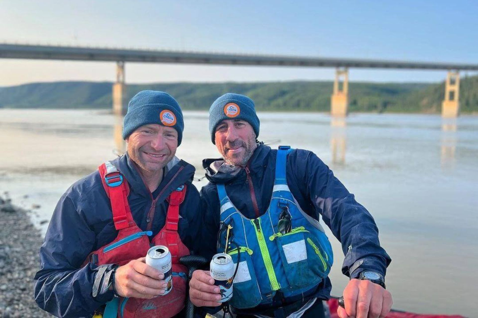 Two canoeists wearing blue beanies, sitting in front of a bridge on a river