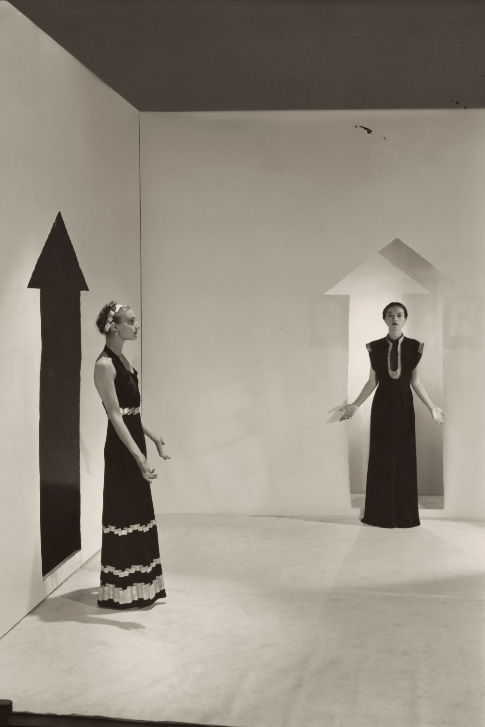 Two models, standing in a white room with arrows painted on walls and wearing dresses by Schiaparelli; Left in a dark crepe with bands of gold braids down the front, over satin pajamas; Right, in a black satin with bands of rippled gold ribbons.