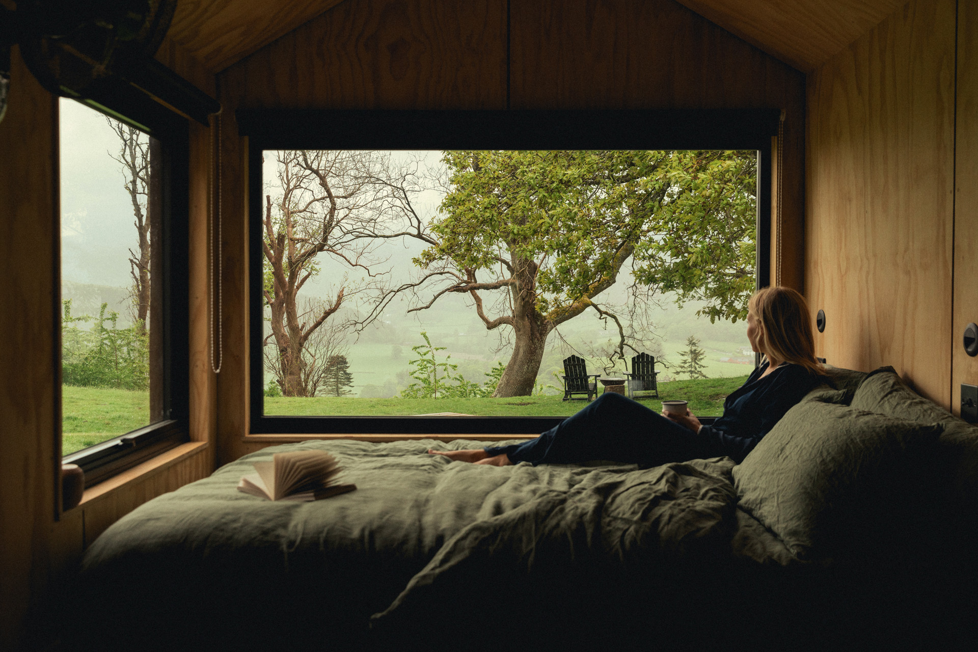 A woman sat on a bed with a huge window looking out onto nature
