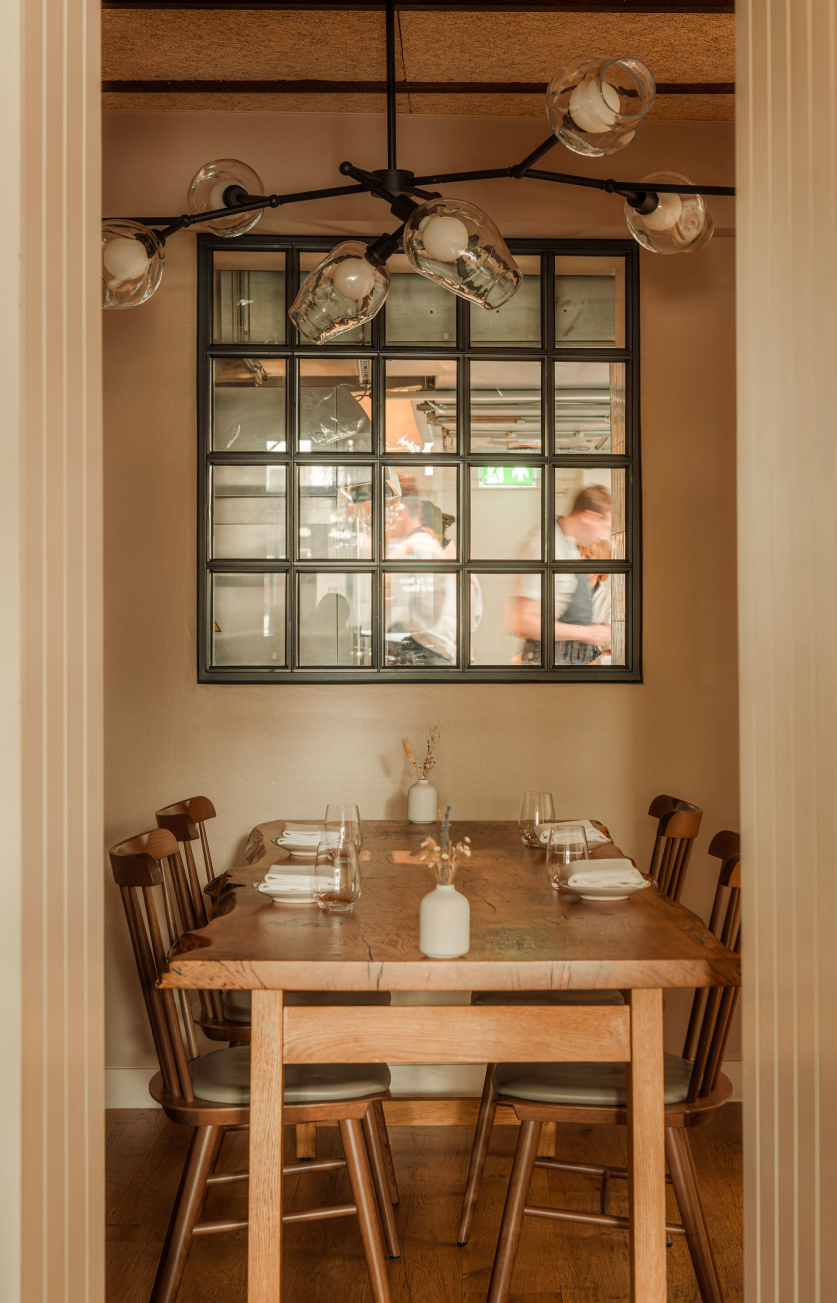 A wooden table and chairs underneath a window looking into a busy professional kitchen