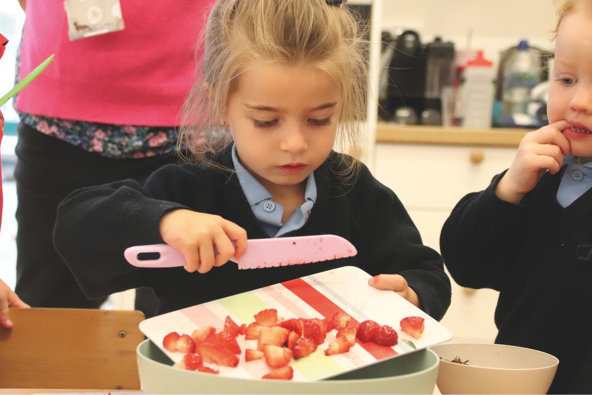 a young blonde girl uses a pink plastic knife to scrape chopped strawberries into a pot