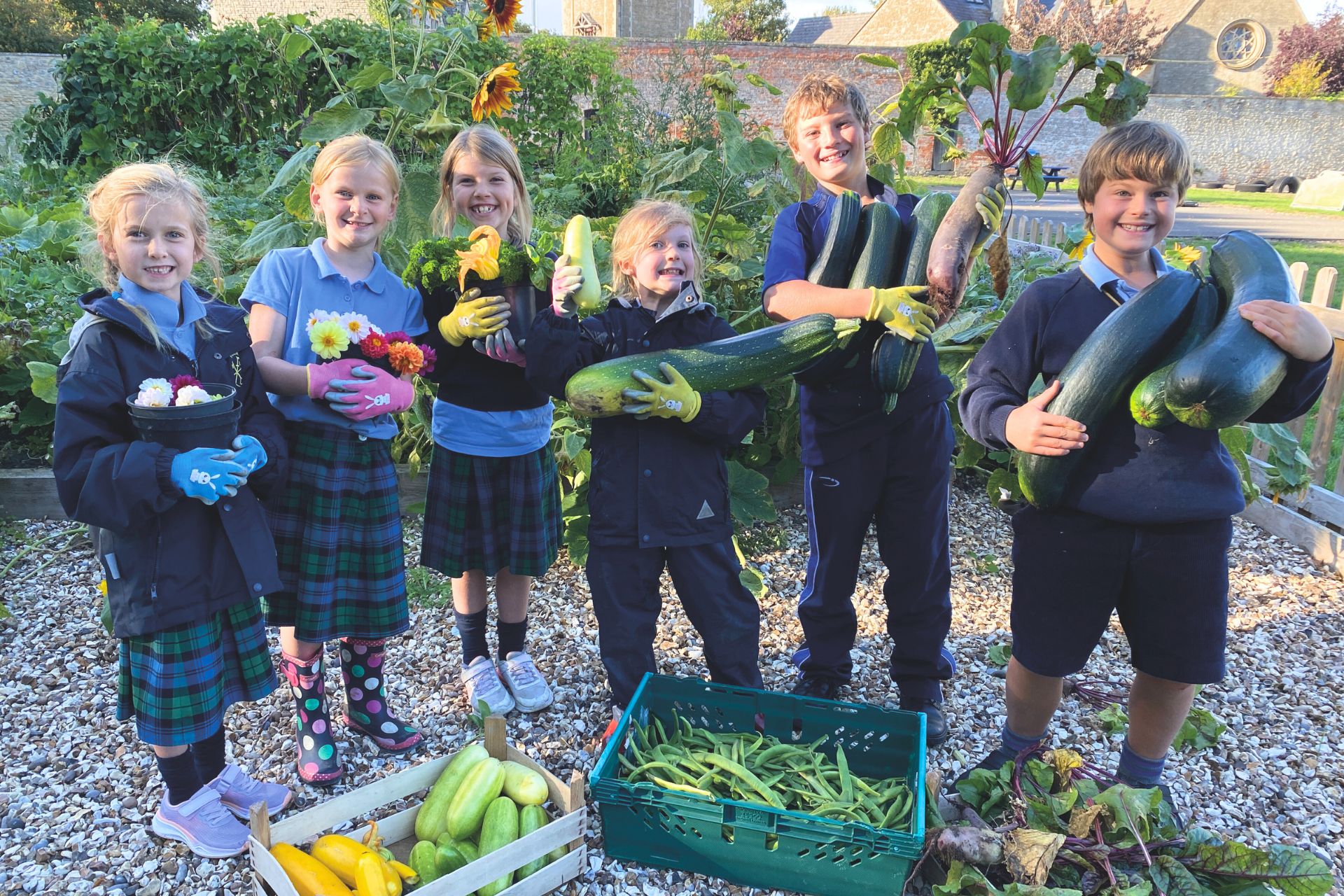 a group of young children show off what they have grown, including very large courgettes, flowers and a crate of pea pods on the ground