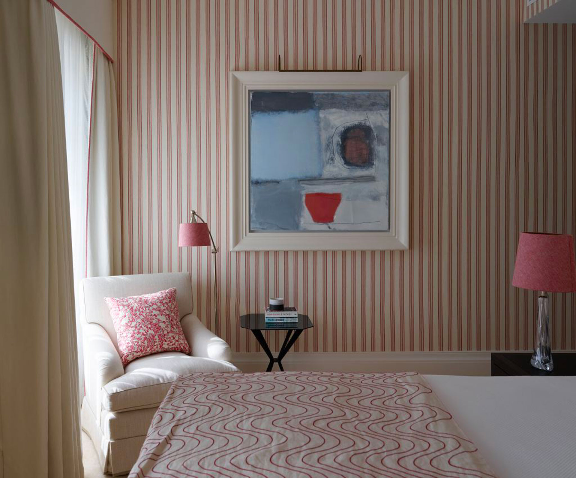 A big painting with a red accent in a room with red striped wallpaper, red quilt, red cushion on chair