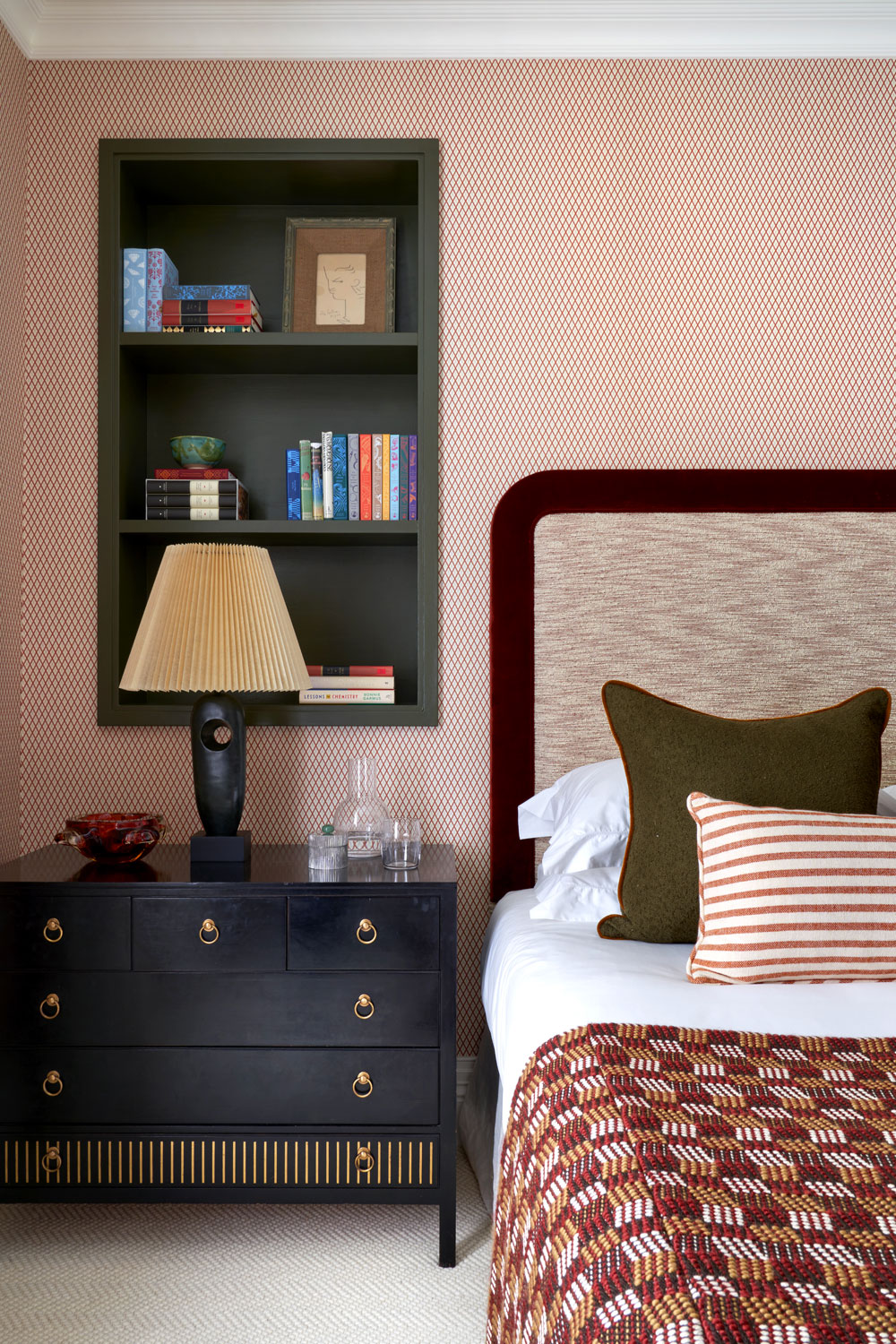 A bed with a red sheet, striped red cushion and green cushion, fabric headboard, next to a side table with a lamp and inset shelving in the wall