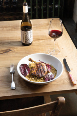A dish of chicken on beans on a wooden table with a glass of red wine