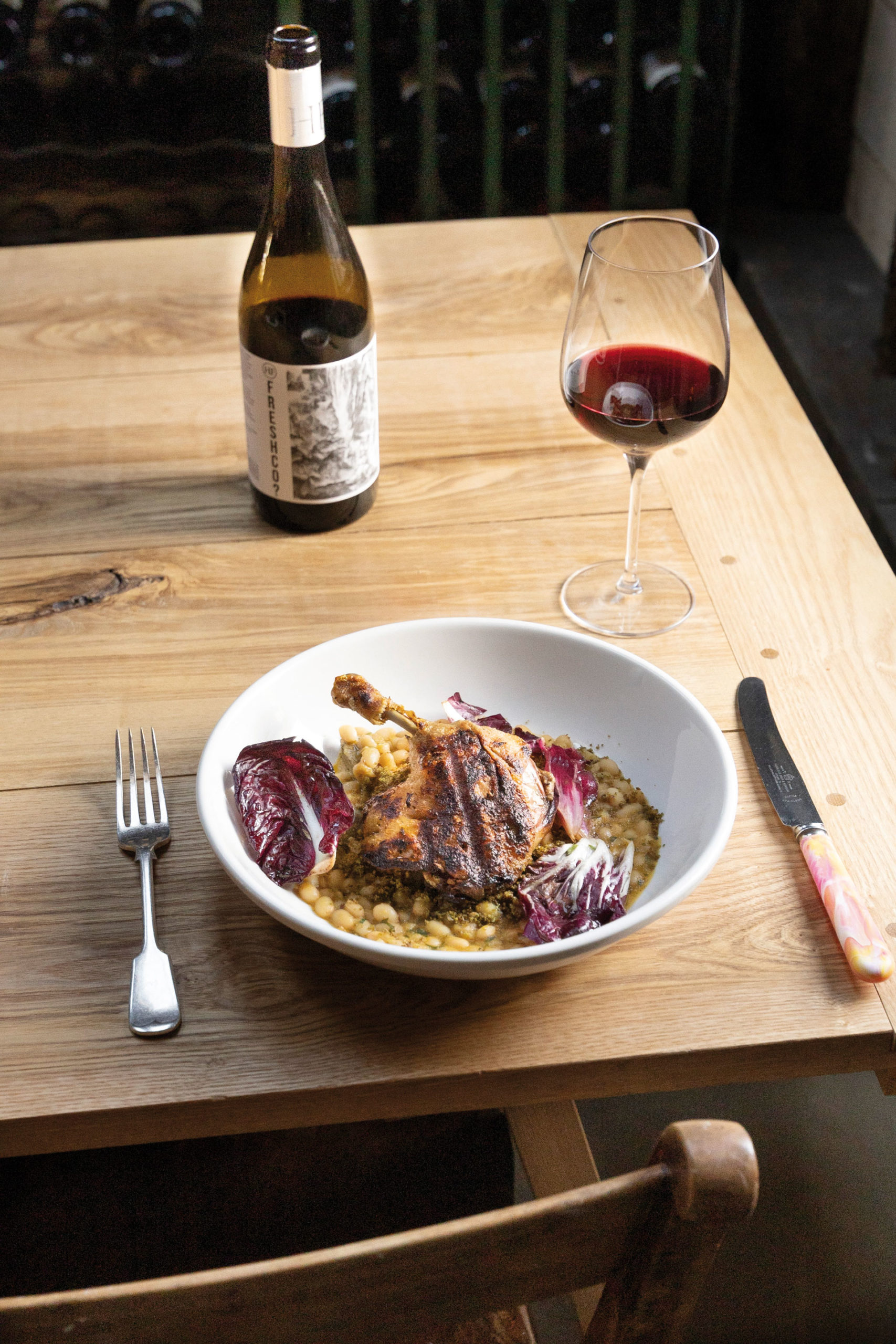 A dish of chicken on beans on a wooden table with a glass of red wine