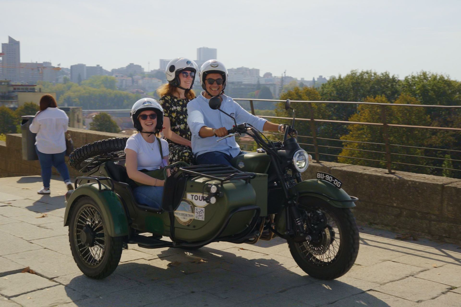 A man riding a motorbike with a woman at the back and another woman to the side in a side car, with a view over Porto in the background