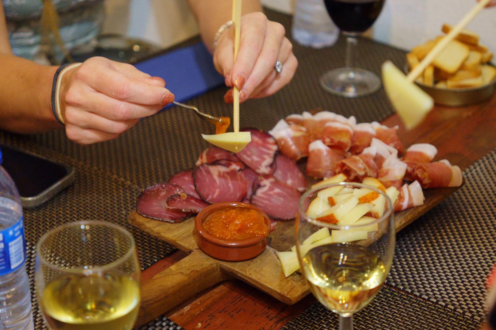 Hands reaching for ham and cheese on a platter with glasses of wine in the foreground
