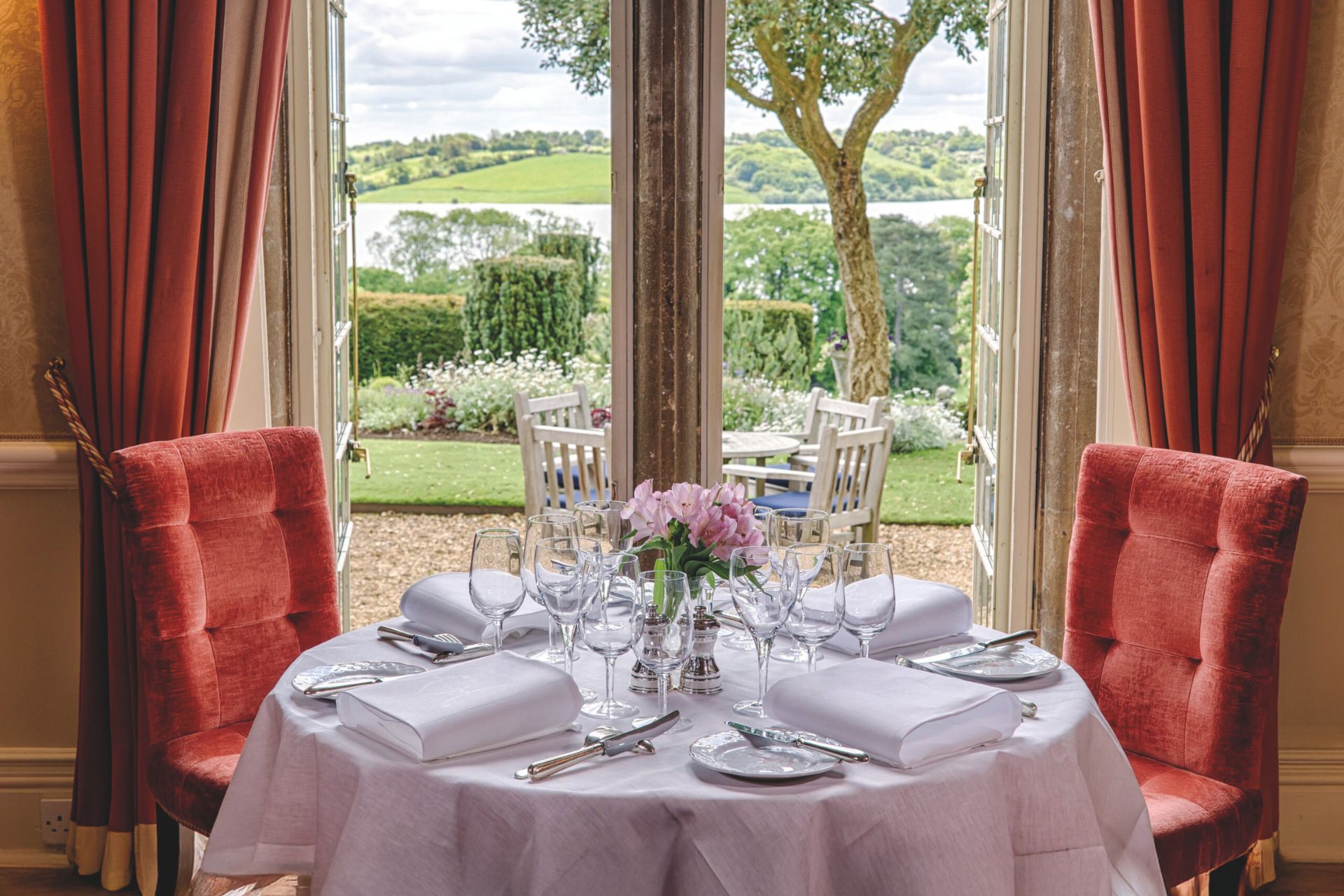 a white cloth dining table and red chairs in front of a view of greenery and water