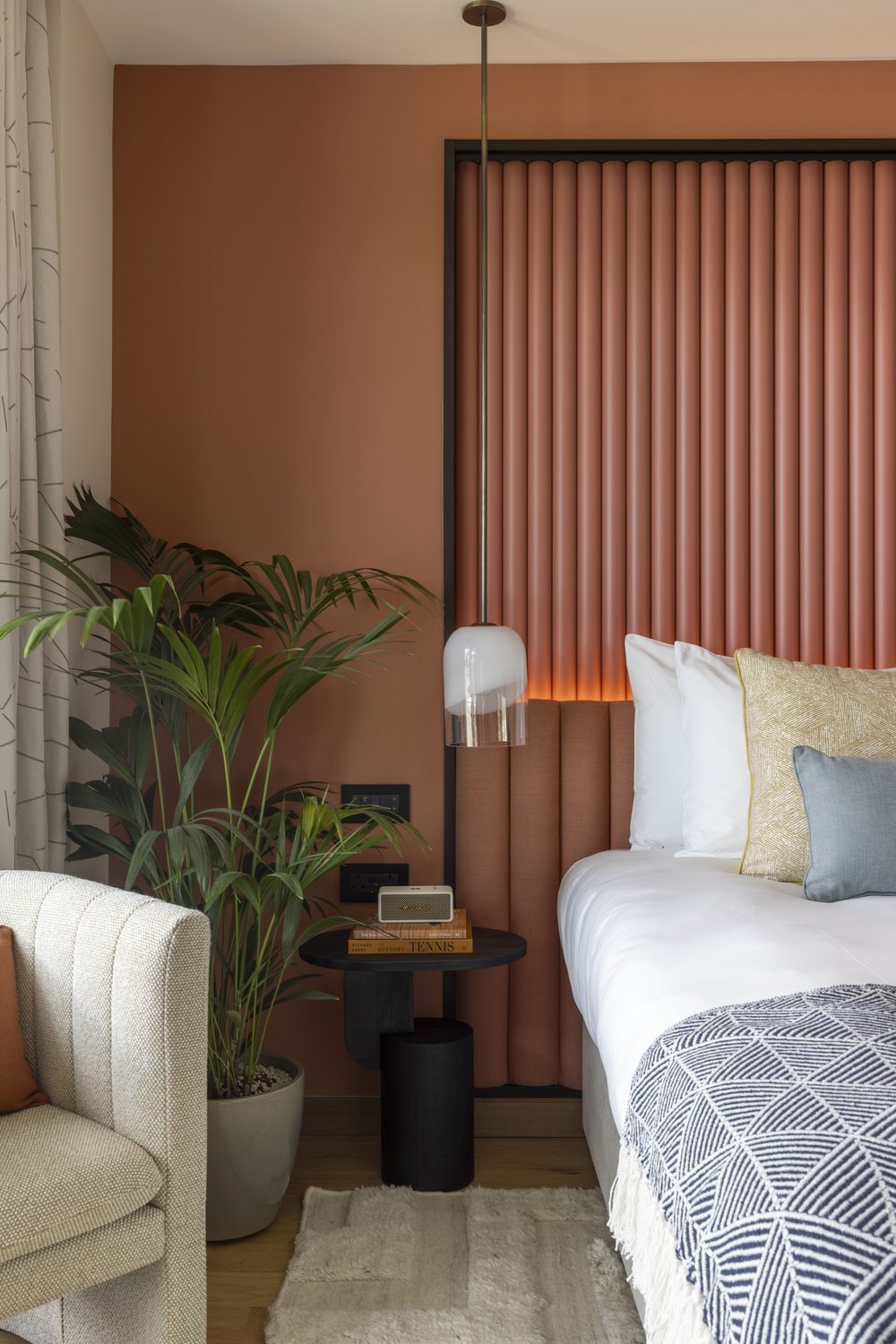 A portrait image of a bed beside a bedside table with a plant and an armchair. The back wall is terracotta-hued with a matching headboard.