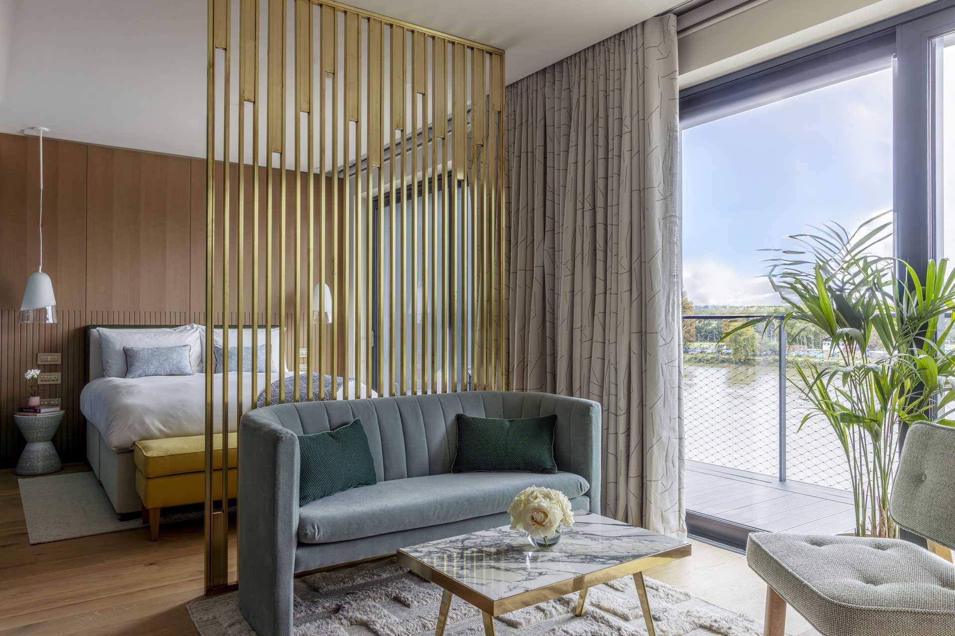 A junior suite at Fulham Pier Hotel with a sofa, coffee table, and bed separated by a room divider, with walk-to-ceiling windows and a balcony