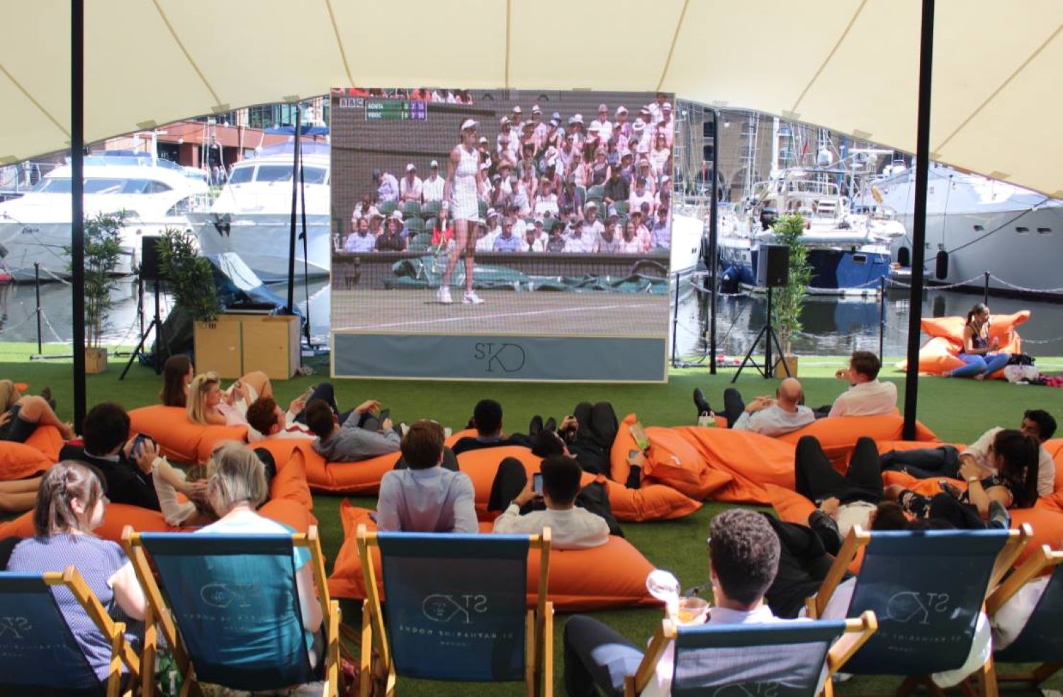 A crowd sat on deck chairs and bean bags under an awning, watching Wimbledon on a big screen.