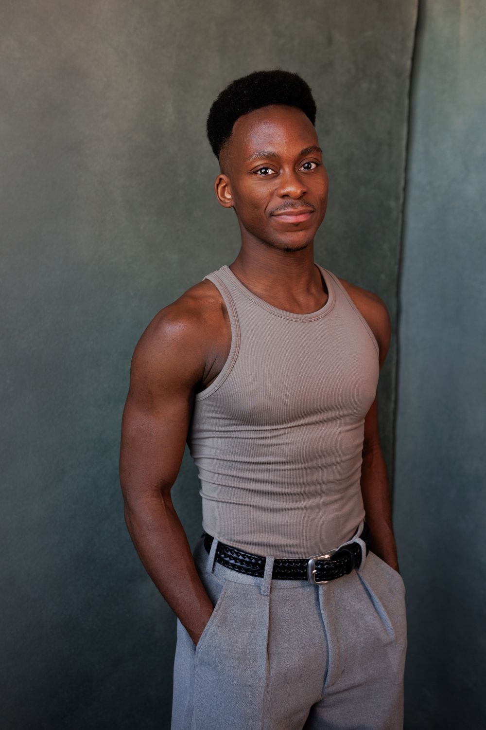 A natural shot of Tyrone wearing a grey singlet and grey trousers, smiling at the camera with his hands in his pockets.