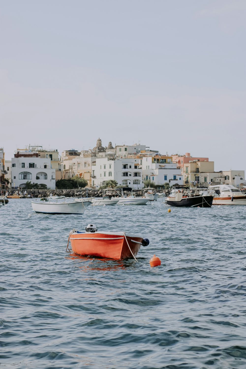 An orange fishing boat in the foreground with whitewashed buildings on the coast of Ischia in the background.