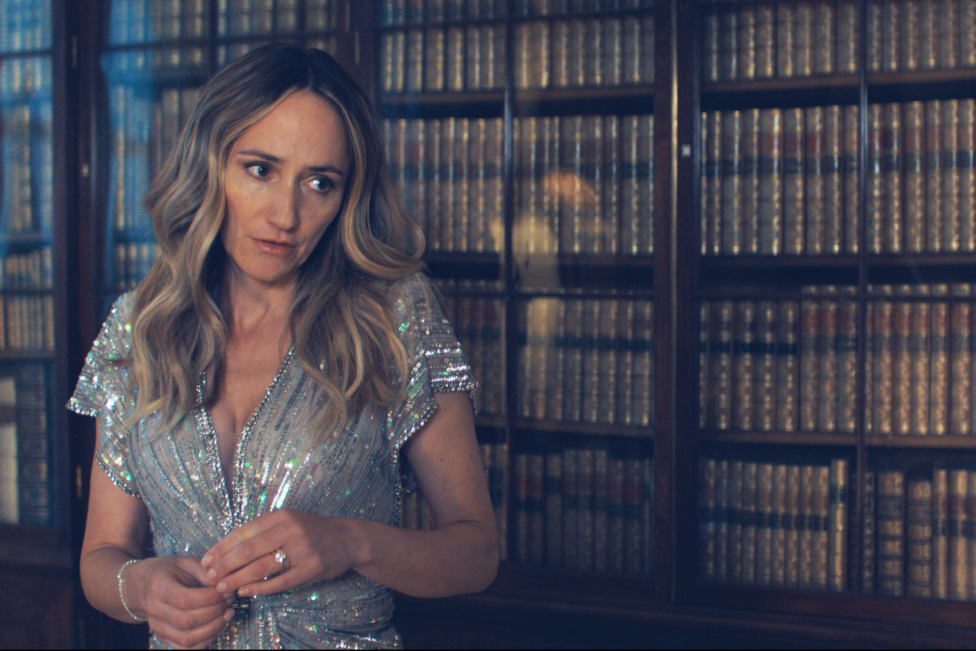 Lydia Leonard in character as Serena in a still from The Party. She stands in a sparkly dress beside bookshelves filled with antique books.