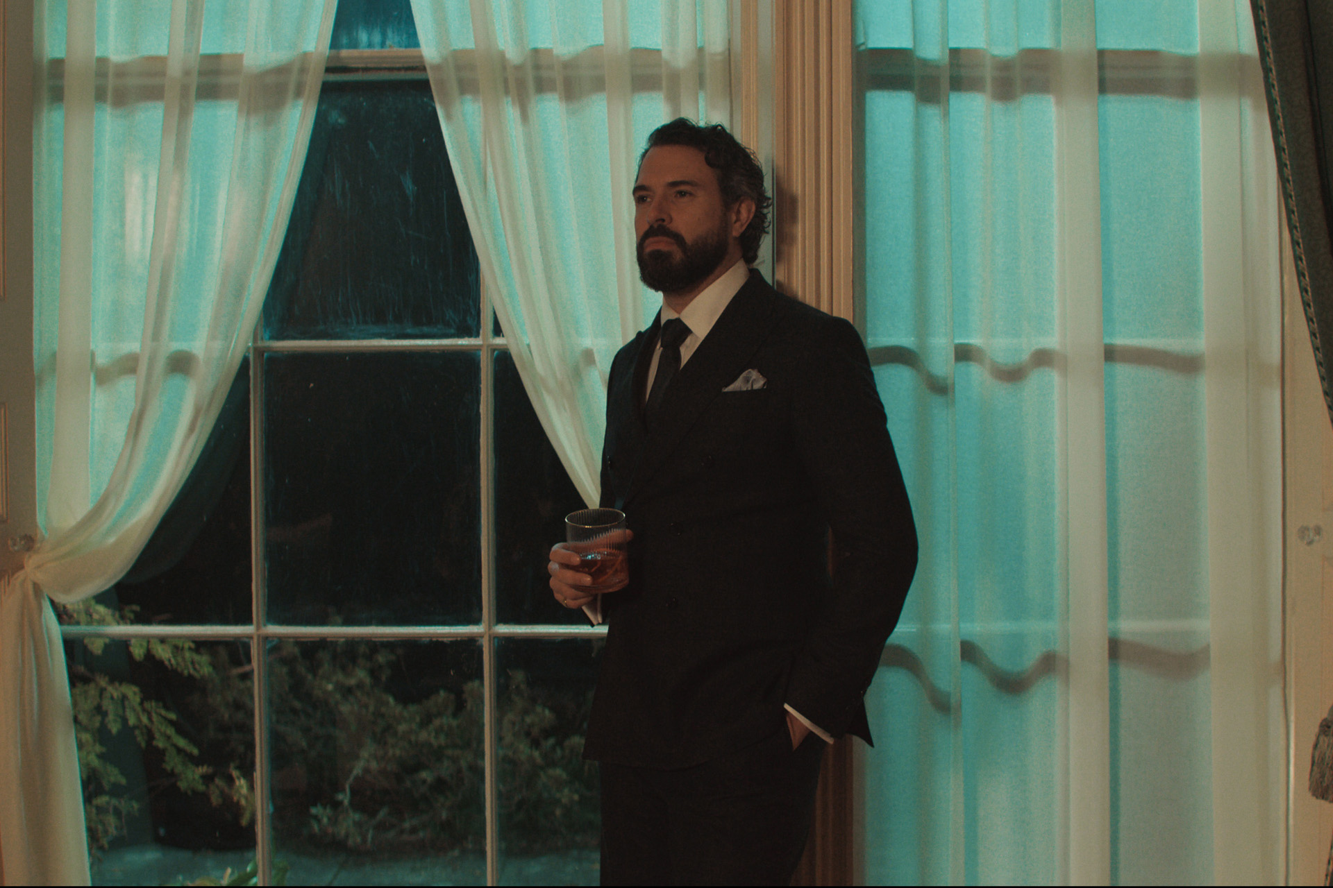 Tom Cullen in character as Ben Fitzmaurice in a still from The Party. He wears a dark suit and stands beside curtained windows with a glass in his hand