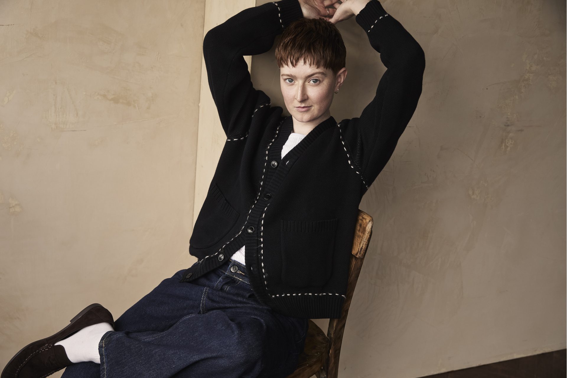 Rex in a cardigan and jeans, leaning back on a chair with their arms raised overhead
