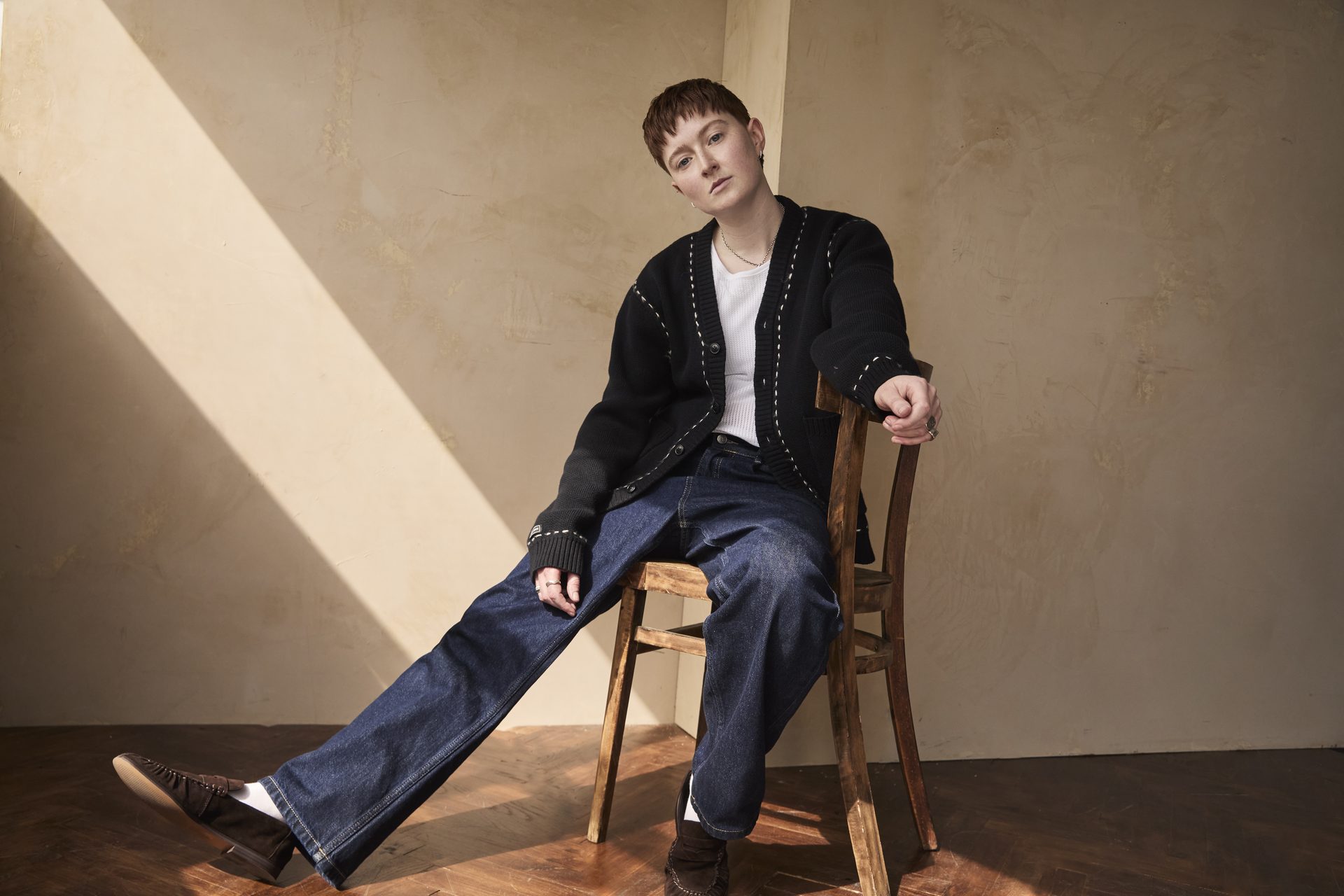 Rex in jeans and a cardigan, sitting sideways on a chair with one leg kicked out to the left