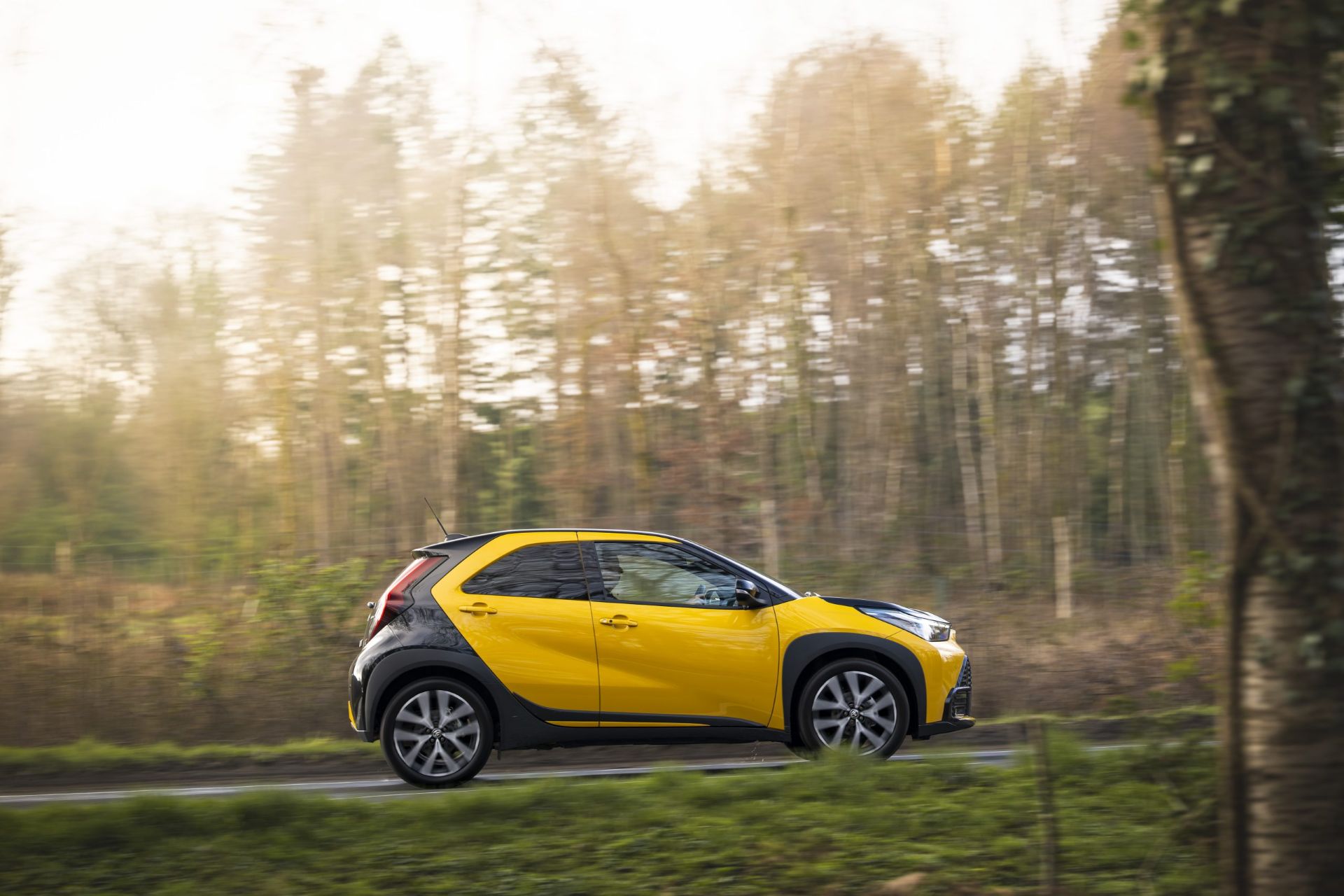 The side of a yellow Toyota Aygo X as it climbs a tree-lined road